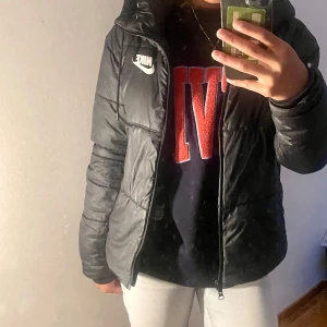 Svart  pufferjacka från Nike - Säljer en svart dunjacka från Nike  Jackan är perfekt för kalla dagar med sin pufferstil och långa ärmar. Den har en dragkedja framtill och en huva för extra värme som är justerbar inga defekter och super bra skick 