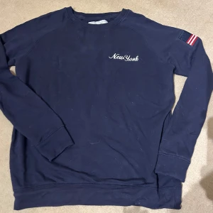 Mörkblå sweatshirt från Lager 175 - Säljer en snygg mörkblå sweatshirt från Lager 175  med texten 'New York' på bröstet. Tröjan har långa ärmar och en liten flaggdetalj på ärmen. Perfekt för en casual look eller en chill dag hemma. 🗽