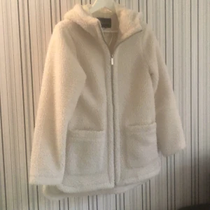 Pälsjacka - Mjuk, mysig jacka från Happy Holly med ett Teddy material. Fodrad, ej avtagbar huva, två praktiska fickor framtil, längd från axel 78 i storlek 36/38. Den känns mer som  XS/S. Jag köpte 2 st, jag säljer den oanvända. 