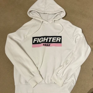 Vit hoodie från Gina Tricot - Säljer en vit hoodie från Gina Tricot med texten 'Fighter 1992' i svart och rosa på framsidan. Den har en klassisk passform med långa ärmar och en mysig huva. Perfekt för en avslappnad stil eller träning. 🖤