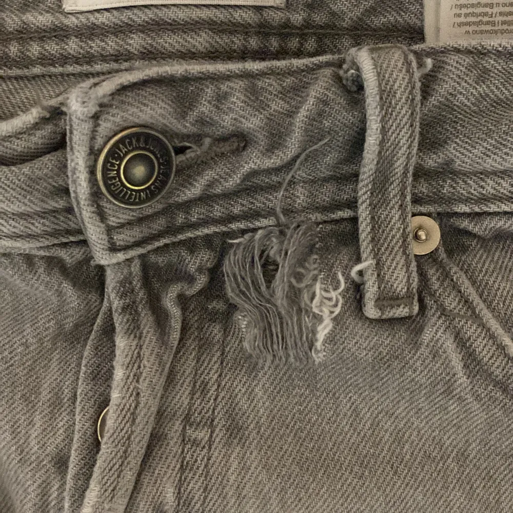 Säljer ett par grå jeans från Jack & Jones. De har en klassisk femficksdesign och en knappgylf. Jeansen är i en ljusgrå nyans och verkar vara i välanvänt skick. Perfekta för en avslappnad vardagslook jeansen har även ett litet hål vid knäppen men de syns inte för man kan ha tröja över!. Jeans & Byxor.