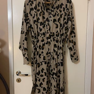 Leopardmönstrad klänning - Säljer en leopardmönstrad klänning i beige och svart i storlek S. Den har långa ärmar och en knytning i midjan för en snygg passform. Perfekt för höst och vår, och funkar lika bra till vardags som till fest. Materialet känns mjukt och bekvämt.