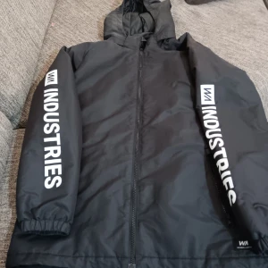 Svart vindjacka från Warp - Säljer en svart vindjacka från Warp med tryck på ärmarna. Jackan har en dragkedja framtill och en huva för extra skydd mot väder. Perfekt för blåsiga dagar och har en cool streetwear-look. Passar bra till både vardag och fritid.