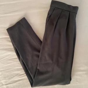 Svarta kostymbyxor - Säljer ett par stilrena svarta kostymbyxor från monki. De har en klassisk design med veck framtill och en elastisk midja för extra komfort. Har textilfiber och lite noppror inget som syns om man inte står nära. Går säkert få bort med maskin.