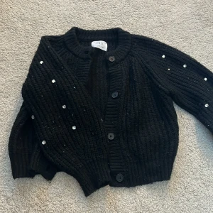Svart stickad kofta från Zara - Säljer en superfin svart stickad kofta från Zara. Den har glittriga detaljer på ärmarna som ger en extra touch. Perfekt för höst och vinter när man vill vara både varm och stilren. Koftan har knappar framtill och är i ett mjukt material som känns skönt mot huden.