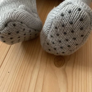 Grå stickade babysockor från Newbie - Säljer ett par supergulliga grå stickade babysockor från Newbie. De har ett sött björnansikte framtill och är fodrade med mjukt vitt foder för extra värme. Sulan har små gummidetaljer för bättre grepp. Perfekta för att hålla små fötter varma under kalla dagar!