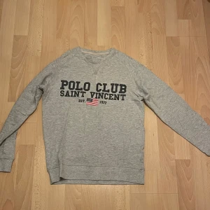 Grå sweatshirt från Polo Club Saint Vincent - Säljer en snygg grå sweatshirt från Polo Club Saint Vincent. Den har ett tryck på framsidan med texten 'POLO CLUB SAINT VINCENT' och en liten amerikansk flagga. Perfekt för en avslappnad stil och passar till både jeans och joggers. Skön och bekväm för vardagsbruk!