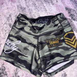 Säljer ett par coola kamouflagefärgade shorts från Anarchy Apparel. De är perfekta för träning och har en tight passform. Shortsen har en grön och svart camo-design med texten 'Whiskey Tango Foxtrot' tryckt på sidan. De är gjorda i ett bekvämt och stretchigt material, perfekt för att röra sig fritt i. Passar perfekt för sommarens alla träningspass!