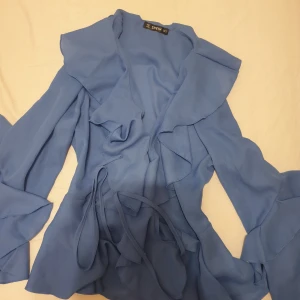 Blus - En blå blus med långa ärmar och volanger. Blusen har en V-ringning och en knäppning framtill. Den är tillverkad i ett tunt, lätt material. Säljer för 70