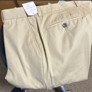 Beige Chinos byxor helt ny med etikett nypris 1295:- - Säljer ett par stilrena beige kostymbyxor i storlek 52. De har en klassisk design med en knapp och dragkedja framtill. Perfekta för både formella tillfällen och kontoret.