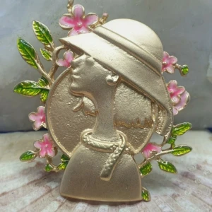 Brosch  - Kvinna i Hatt / Modelejon  - En brosch föreställande en kvinna, storlek ca 4.5x4cm.  Guldfärgad metall med vacker ytstruktur och emaljering bl.a Vacker brosch.