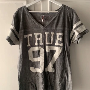 Grå t-shirt med tryck - Säljer en grå t-shirt med  tryck 'TRUE 97' på framsidan och 'LOVE' i rosa på baksidan. Den har en v-ringad hals och korta ärmar med vita ränder. Perfekt för en avslappnad stil! Använd, men i fint skick. 