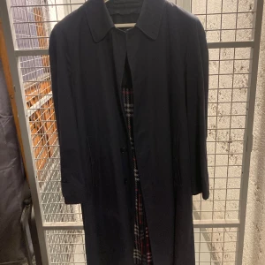 Mörkblå kappa från Burberry - Säljer en elegant mörkblå kappa från Burberry. Den har en klassisk krage och knäppning framtill. Insidan är fodrad med ett snyggt rutigt mönster i svart, vitt och rött i ull. Perfekt för höst och vinter!