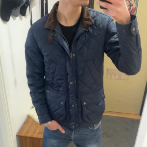 Blå quiltad jacka från Ralph Lauren - Säljer en snygg jacka från Ralph Lauren. Den har en brun krage i manchester och stängs med både dragkedja och knappar. Perfekt för höst och vår med sin stilrena design och praktiska fickor. Jackan är i nyskick och passar perfekt för en cas