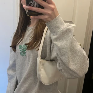 Vit axelväska med dragkedja - Säljer en stilren vit axelväska i syntetmaterial. Väskan har en praktisk dragkedja och är perfekt för vardagsbruk. Den är i bra skick och passar till de flesta outfits. Väskan kommer tvättas innan så fläckar på bandet försvinner!