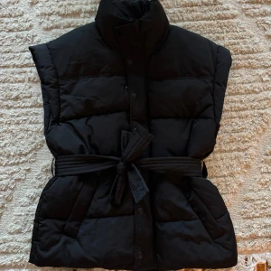 Svart dunväst från Gina Tricot - Säljer en snygg och stilren svart dunväst från Gina Tricot. Västen har en hög krage och knäpps med knappar framtill. Den har ett bälte i midjan för en mer figurnära passform. Perfekt för höst och vinter!