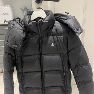 Svart pufferjacka från Calvin Klein - Säljer en snygg svart pufferjacka från Calvin Klein Jeans. Jackan är perfekt för kalla dagar med sin vadderade design och har en praktisk dragkedja framtill. Den har en avtagbar huva och två fickor med dragkedja. Kostar 2399 ny.