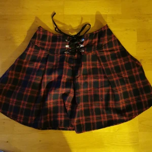 Röd/svartrutig kort kjol  - Röd/svartrutig kort kjol vidare modell i stl M med snörning fram och dragkedja på sidan.  