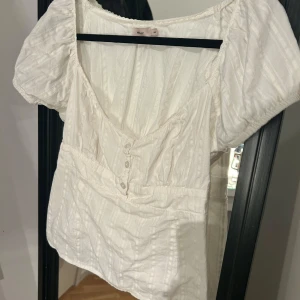 Vit topp  - En super gullig topp från Nelly by Nelly i storlek 40. Den är använd Max 3 gånger och har inga defekter. Det finns en extra knapp på lappen på sidan ifall nån av knapparna vid bysten skulle trilla av. 