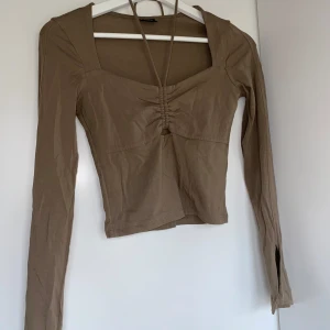 Beige långärmad topp med knytband - Säljer en snygg beige topp med långa ärmar och en cool knytning framtill. Toppen har en tight passform och är perfekt för höst och vår. Den har en liten slits vid ärmsluten som ger en extra detalj.
