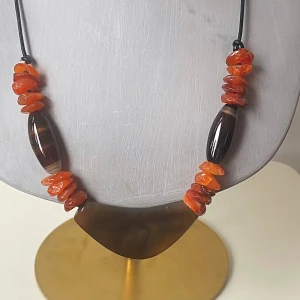Halsband med orange och bruna stenar - Säljer ett unikt halsband med stora, orange och bruna agatstenar. Det har en cool design med en svart snodd som håller ihop de färgglada stenarna. Perfekt för att ge en bohemisk touch till din outfit!