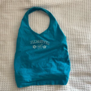 Turkos halterneck topp med tryck - Säljer en snygg turkos halterneck topp med texten 'Vancouver' och blommigt tryck på framsidan. Toppen har en knytning i ryggen och är perfekt för sommardagar. Den är i bra skick och superbekväm att ha på sig!