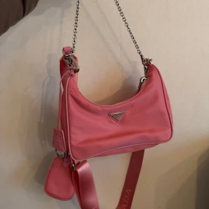 Rosa axelväska  - Snygg rosa axelväska med kedjehandtag och justerbar axelrem. Väskan har en dragkedja och en liten avtagbar väska som hänger på remmen. Perfekt för att ge din outfit en färgklick!