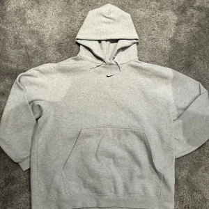 Grå hoodie från Nike - Säljer en klassisk grå hoodie från Nike med en liten svart logga på bröstet. Den har en stor ficka framtill och en justerbar huva med dragsko. Perfekt för en avslappnad stil.