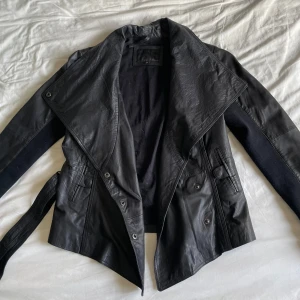 Svart skinnjacka med bälte - Snygg svart skinnjacka med bälte och knappar. Jackan har en cool asymmetrisk design och långa ärmar. Knappt andvänd, av märket frontrow living