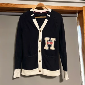 Mörkblå kofta med vita muddar  - Snygg svart kofta från Lula med vita detaljer och ett broderat 'H' med texten 'New York' på bröstet. Koftan har knappar framtill och två fickor. Perfekt för en stilren look.