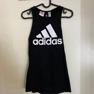 Svart linnetopp från Adidas st 164 -XS - Säljer en svart linnetopp från Adidas med det klassiska vita logotyptrycket på framsidan. Perfekt för träning eller en avslappnad dag. Toppen är ärmlös och har en bekväm passform.