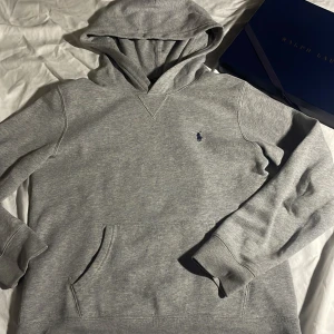 Grå hoodie från Ralph Lauren - Säljer en klassisk grå hoodie från Ralph Lauren med en liten broderad logga på bröstet. Tröjan har en bekväm passform med långa ärmar och en praktisk magficka. Perfekt för en avslappnad stil.