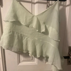 Ljusgrön topp från Gina Tricot - Säljer en ljusgrön topp från Gina Tricot med volangdetaljer. Toppen har en asymmetrisk design med en axelbandslös stil. Perfekt för sommarens varma dagar.