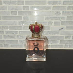 Dolce & Gabbana Q Eau de Parfum - Säljer en lyxig parfym från Dolce & Gabbana med en elegant flaska. Flaskan är fyrkantig med en kunglig krona som lock i rött och guld. Parfymen har en sofistikerad doft och kommer i en 50 ml flaska.