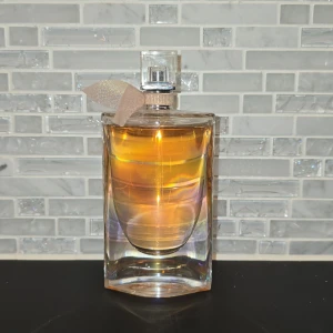 La Vie Est Belle Eau de Parfum - Elegant parfymflaska från Lancôme med en vacker, transparent design och en subtil rosa vätska. Flaskan har en fyrkantig kork och en dekorativ rosett vid halsen. Doften är känd för sin blommiga och fruktiga karaktär, perfekt för den som vill känna sig lyxig och självsäker.