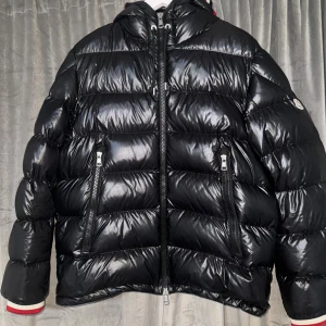  Moncler - Säljer en snygg svart pufferjacka från Moncler med glansigt material och coola detaljer. Jackan har dragkedja och långa ärmar med ribbade muddar i rött, vitt och blått. Perfekt för kyliga dagar med sin quiltade design och huva. Storlek 5 = XL 