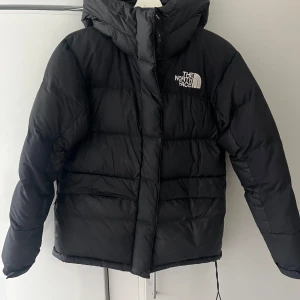 Svart dunjacka från The North Face - Säljer en svart dunjacka i storlek S från The North Face. Jackan har en pufferdesign med lång ärm och en praktisk huva. Logotypen är tryckt på bröstet och ryggen.