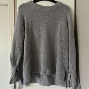 Grå stickad tröja med knytärmar - Mysig grå stickad tröja med långa ärmar och knytning vid ärmsluten. 14% ull