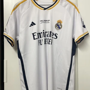 Real Madrid fotbollströja - Säljer en vit Real Madrid fotbollströja från Adidas med korta ärmar. Tröjan har gula och svarta detaljer samt klubbens emblem på bröstet. Texten 'FINAL LONDON 2024' är tryckt på framsidan. Köpte tröjan i London för champions league och är använd minst 1 gång. Storlek L men det är slimfit så det passar M Perfekt för fans av Real Madrid!