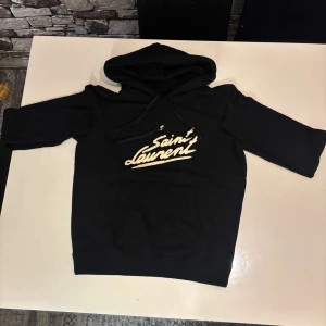 Svart hoodie från Saint Laurent - Snygg svart hoodie från Saint Laurent med vit logga på framsidan. Tröjan har en klassisk design med huva och dragsko samt långa ärmar. Perfekt för en stilren och bekväm look.