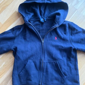 Mörkblå hoodie från Ralph Lauren - Säljer en snygg mörkblå hoodie från Ralph Lauren med dragkedja och huva. Den har en liten röd logga på bröstet och är perfekt för en avslappnad stil. Passar bra till både jeans och joggers. Tröjan är köpt på KidsBrandStore för 1200kr. Spris kan diskuteras:)