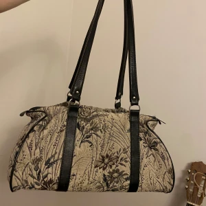 Axelväska med blommigt mönster - Säljer en unik axelväska med blommigt mönster i beige och grått. Väskan har svarta handtag och detaljer. Väskan är ganska stor, ca 40x15cm. Skriv om du har fler frågor💕