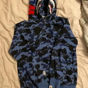 Blå camo hoodie från A Bathing Ape - Säljer en blue camo hoodie från A Bathing Ape med hajmotiv på huvan. Hoodien har dragkedja och är tillverkad i bomull. Perfekt för dig som vill sticka ut med en unik stil. 🦈 Hör av er vid frågor!🙂💛