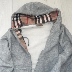 Grå hoodie från Burberry - Säljer en stilren grå hoodie från Burberry med klassiskt rutigt mönster i huvan. Tröjan har dragkedja och är perfekt för en avslappnad look. Passar bra till både vardag och chill.