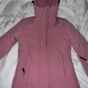 Rosa dunjacka från cross sportswear - Säljer en snygg rosa dunjacka från cross sportswear. Perfekt för skidåkning med en ficka för liftkort. Jackan har en justerbar huva och dragkedja framtill. Perfekt för kalla dagar med sin vadderade design. Den har långa ärmar och praktiska fickor med dragkedjor. Den är i storlek small men skulle säga lite åt det mindre hållet.