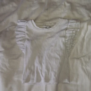 Vit ribbad topp från Zara - Säljer en söt vit ribbad topp från Zara med volangdetaljer på ärmarna. Perfekt för en stilren look. 
