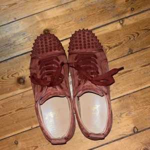 Röda sneakers med nitar från Christian Louboutin - Snygga unika sneakers i mocka från Christian Louboutin med nitar på tån och klassisk röd sula. Skorna har snörning och en stilren design som sticker ut. Låda tillkommer ej.