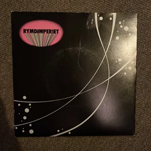 Rymdimperiet - Vad Pojkar Vill Ha / Bikinipop 7” vinylsingel - Vinylsingel från Rymdimperiet med låtarna 'Vad Pojkar Vill Ha' och 'Bikinipop'. Utgiven 1981 av Mistlur. Omslaget är svart med vita linjer och rosa text. Perfekt för samlare av svensk punk och rock.