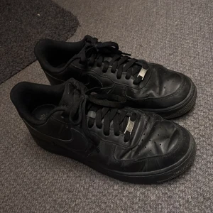 Svarta Nike Air Force sneakers - Säljer ett par klassiska svarta Nike Air Force sneakers. Skorna har en stilren design med snörning och perforerade detaljer på ovansidan för bättre ventilation. Perfekta för en casual look.