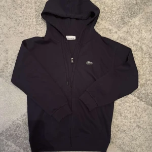 Mörkblå hoodie från Lacoste - Säljer en stilren mörkblå hoodie från Lacoste med dragkedja och den klassiska krokodilloggan. 